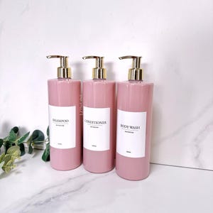 May include: Three pink plastic bottles with gold pumps and white labels. The labels read "Shampoo Bathroom 500ml", "Conditioner Bathroom 500ml", and "Body Wash Bathroom 500ml".