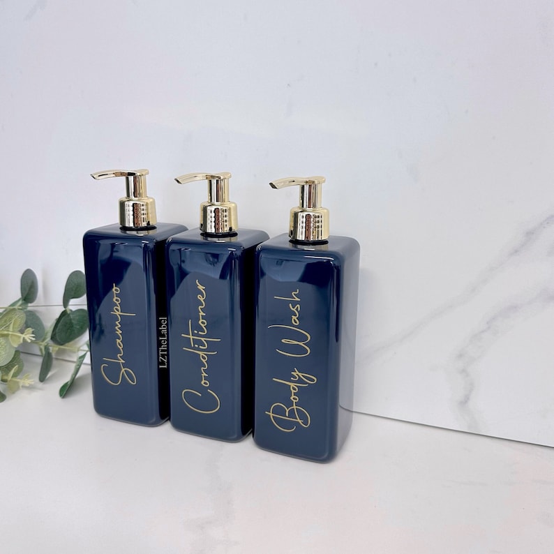 K&ouml;nnte beinhalten: Drei dunkelblaue Glaspumpflaschen mit goldenen Aufs&auml;tzen und goldenen Beschriftungen. Die Flaschen sind mit "Shampoo", "Conditioner" und "Duschgel" beschriftet.