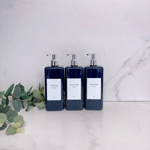 Pode incluir: Três garrafas retangulares de cor azul escuro com bombas prateadas e etiquetas brancas. As etiquetas dizem "Shampoo", "Condicionador" e "Gel de banho".