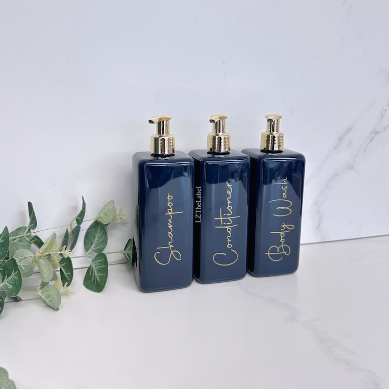 K&ouml;nnte beinhalten: Drei dunkelblaue Pumpflaschen mit goldenen Verschl&uuml;ssen und Etiketten. Die Etiketten zeigen "Shampoo", "Conditioner" und "Duschgel" in goldener Schrift.
