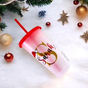 May include: A white, frosted, reusable cup with a red lid and a red and white Santa hat design. The cup has a gold letter "S" with the name "Sarah" written in cursive. The cup is on a white background with Christmas decorations.