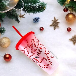 May include: A white tumbler with a red lid and a straw. The tumbler has a design of candy canes and the word "Grace" written in cursive. The tumbler is on a white surface with a Christmas tree and decorations in the background.