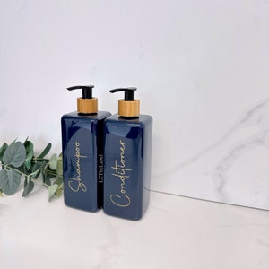K&ouml;nnte beinhalten: Zwei blaue Pumpflaschen mit Bambusdeckeln und goldener Beschriftung. Die Flasche links tr&auml;gt die Aufschrift "Shampoo" und die Flasche rechts "Conditioner".