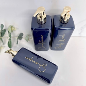 K&ouml;nnte beinhalten: Drei marineblaue Pumpflaschen mit goldenen Verschl&uuml;ssen und Etiketten. Die Etiketten sind in goldener Schrift geschrieben und lauten "Shampoo", "Conditioner" und "Duschgel".