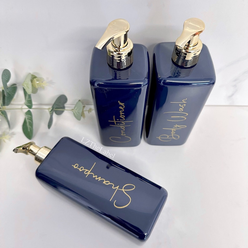 K&ouml;nnte beinhalten: Drei marineblaue Pumpflaschen mit goldenen Verschl&uuml;ssen und Etiketten. Die Etiketten sind in goldener Schrift geschrieben und lauten "Shampoo", "Conditioner" und "Duschgel".