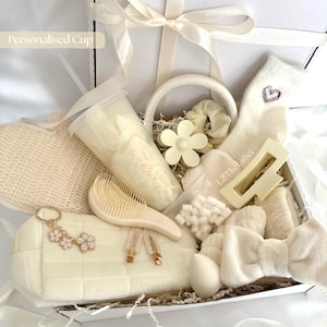 May include: A gift box containing a cream-colored tumbler with a straw, a quilted pouch with a floral keychain, a hairbrush, a flower-shaped hair clip, a headband with a bow, fuzzy socks with a heart, a scrunchie, and hair accessories.