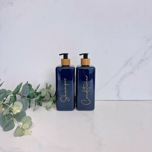 K&ouml;nnte beinhalten: Zwei dunkelblaue Plastikflaschen mit Bambuspumpen und goldenen Etiketten. Die Etiketten lauten "Shampoo" und "Conditioner".