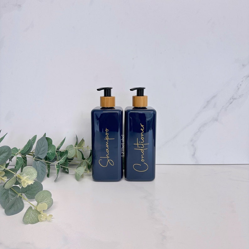 K&ouml;nnte beinhalten: Zwei dunkelblaue Plastikflaschen mit Bambuspumpen und goldenen Etiketten. Die Etiketten lauten "Shampoo" und "Conditioner".