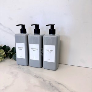 Peut inclure: Trois flacons en plastique gris avec des pompes noires et des étiquettes blanches. Les étiquettes indiquent "Shampooing", "Après-shampoing" et "Gel douche".