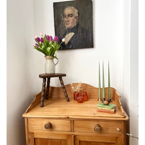 May include: An antique interior with a wooden cabinet, a small wooden stool, and a portrait. A vase of purple and pink tulips sits on the stool. Three green candles and small orange vases are on the cabinet. Website address visible.