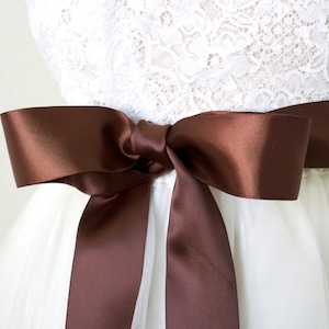 May include: A brown satin ribbon tied in a bow around a white dress. The ribbon is approximately 5 cm wide.