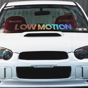 Puede incluir: Un Subaru Impreza blanco con una calcomanía "LOW MOTION" arcoíris en el parabrisas.