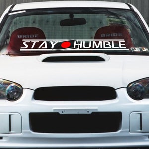 Puede incluir: Coche blanco con una pancarta de parabrisas "STAY HUMBLE" negra y roja.
