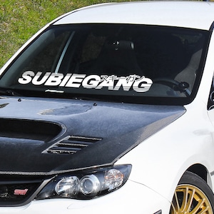 Puede incluir: Subaru blanco con un capó de fibra de carbono negro y una pancarta en el parabrisas que dice "SUBIEGANG" en blanco con un gráfico de caracteres japoneses.