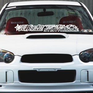 Puede incluir: Coche blanco con una pancarta en el parabrisas que dice "STAY HUMBLE" en texto blanco y caracteres japoneses. Se ven asientos BRIDE rojos a través del parabrisas.