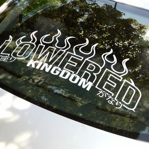 Op de afbeelding: Een witte vinyl sticker met de tekst "LOWERED KINGDOM" in een gestileerd lettertype met vlammen aan beide kanten. De tekst "かならず" is ook in Japanse karakters opgenomen.
