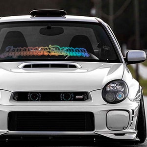 Könnte beinhalten: Ein weißer Subaru Impreza mit einem regenbogenfarbenen "Japanese Demon" Aufkleber auf der Windschutzscheibe.