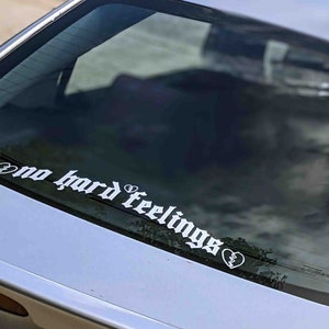 May include: White vinyl car decal with the phrase "no hard feelings" in a gothic font, with broken heart graphics on each end. The decal is applied to the rear window of a silver car.