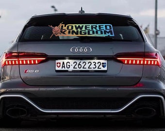 Pode incluir: Um carro cinzento com um autocolante "Lowered Kingdom" no vidro traseiro. O autocolante apresenta um gráfico e texto em cores degradê. O carro tem luzes traseiras vermelhas e uma chapa de matrícula com o texto "AG-262232".