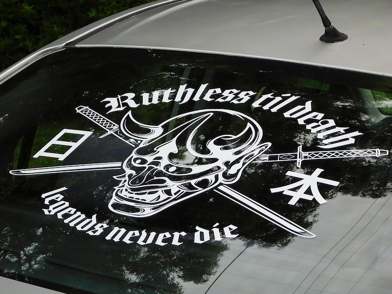 Op de afbeelding: Een witte vinyl sticker met een zwart-witte illustratie van een Japans demonenmasker met twee gekruiste zwaarden erachter. De tekst "Ruthless til death" staat boven het masker en "legends never die" staat onder het masker. De Japanse karakters "日" en "本" staan aan weerszijden van het masker.