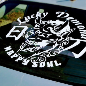 Könnte beinhalten: Ein weißer Vinyl-Aufkleber mit einer Schwarzweiß-Illustration eines Dämons mit dem Text "Lucky Demon Happy Soul" drumherum.