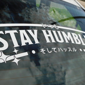 Może przedstawiać: Biała naklejka winylowa z tekstem "Stay Humble" w pogrubionym kroju. Naklejka zawiera również dwie małe gwiazdki i japoński tekst "そしてハッスル", który tłumaczy się jako "i hustle".