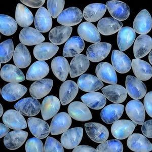 Könnte beinhalten: Eine Sammlung birnenförmiger Mondstein-Cabochons, jeder mit einem einzigartigen irisierenden Schimmer. Die Steine sind milchig weiß mit blauen und grünen Schimmern.