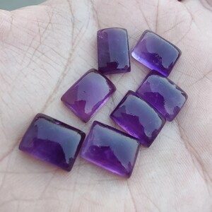Puede incluir: Siete piedras preciosas de amatista rectangulares pulidas con esquinas redondeadas. Las gemas son de un púrpura profundo y translúcido, que refleja la luz. Las piedras se exhiben sobre una superficie clara, mostrando su color y claridad.
