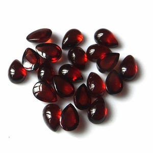 Può includere: Una collezione di 18 cabochon di pietre preziose rosse a forma di goccia. I cabochon sono lisci e lucidi, di un colore rosso intenso.