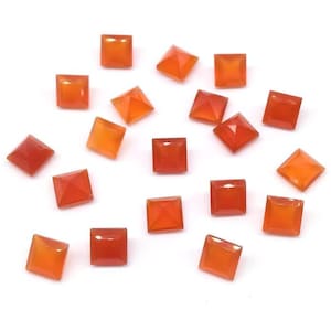 Könnte beinhalten: Eine Sammlung quadratisch geschliffener, orangefarbener Edelsteine. Die Edelsteine sind facettiert, mit einer Mischung aus flachen und abgewinkelten Oberflächen, und sind vor einem weißen Hintergrund verstreut. Die Farbtöne der Edelsteine variieren von hellorange bis tiefrot-orange.