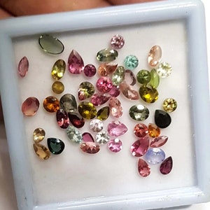 May include: A collection of colourful gemstones in various shapes and sizes, including green, pink, red, and yellow. The gemstones are displayed in a white box with a light grey border.