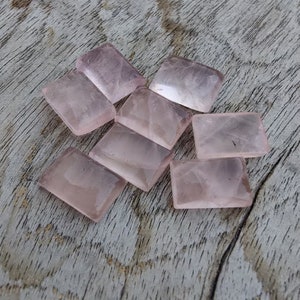 May include: Eight rectangular pink rose quartz stones, with a smooth, polished finish, are scattered on a light brown wooden surface.