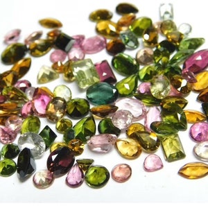May include: A collection of loose gemstones in various shapes and sizes, including green, pink, brown, and black. The gemstones are scattered on a white surface.