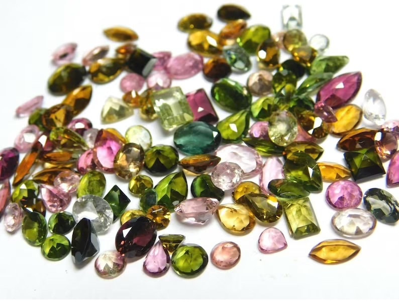 May include: A collection of loose gemstones in various shapes and sizes, including green, pink, brown, and black. The gemstones are scattered on a white surface.