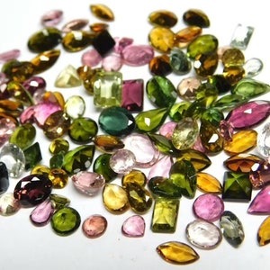 May include: A collection of loose gemstones in various shapes and sizes, including green, pink, brown, and clear stones. The stones are arranged on a white surface.