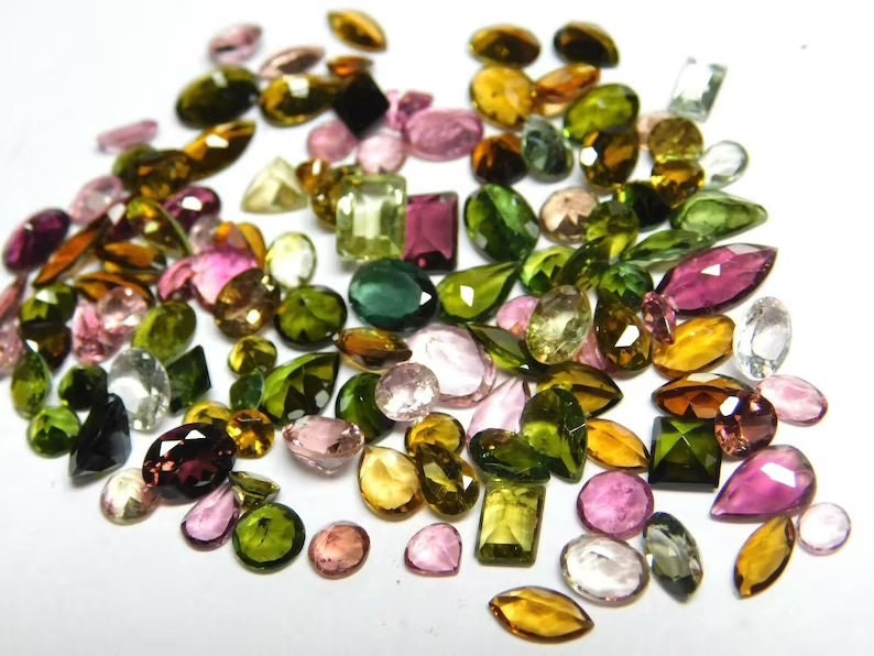 May include: A collection of loose gemstones in various shapes and sizes, including green, pink, brown, and clear stones. The stones are arranged on a white surface.