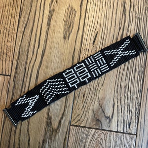 May include: Black and white beaded bracelet with a geometric pattern. The bracelet is made of small black beads with white beads forming a repeating design.
