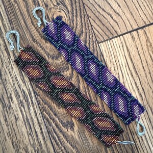 May include: Two beaded bracelets with silver clasps. One bracelet is purple and black with a geometric pattern. The other bracelet is brown, green, and gold with a wavy pattern.