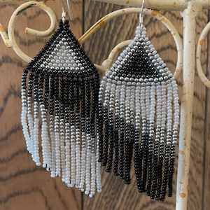 May include: A pair of black and white beaded earrings with a triangular shape at the top and a fringe of beads hanging down. The earrings are ombre, with the top half being black and the bottom half being white.