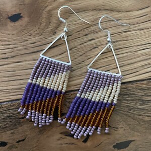 Puede incluir: Un par de pendientes colgantes con un marco triangular de plata y flecos de cuentas. Los flecos están hechos de cuentas moradas, marrones, crema y blancas.