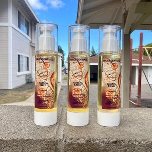 May include: Three bottles of Golden Elixir facial oil are displayed on a concrete ledge. The clear bottles have white pumps and bases, with labels featuring orange slices and the product name. The background includes a building and a blue sky.