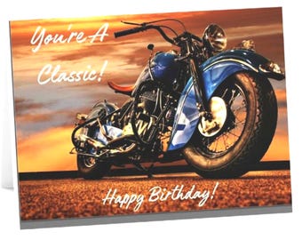 May include: A greeting card featuring a blue vintage motorcycle parked on a road at sunset. The text on the card reads "You're A Classic!" in the upper left and "Happy Birthday!" at the bottom. The motorcycle has a large front wheel and chrome details.