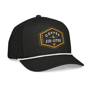 May include: Black baseball cap with a white rope detail and a flat peak. The cap features a gold and white graphic that reads "Coffee & Jiu-Jitsu" in a hexagon shape. The cap has a button on top.