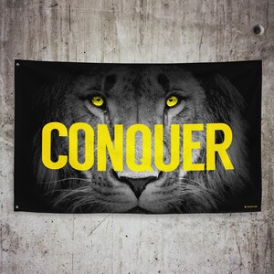 May include: Black and white image of a lion's face with yellow eyes. The word "CONQUER" is written in large yellow letters across the lion's face.