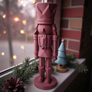 Op de afbeelding: Een stoffig roze houten notenkrakerfiguur met een kroon, staand op een ronde basis. Een kleine blauwe kerstboom en een dennenappel staan ook op de vensterbank. De achtergrond omvat een bakstenen muur en vage lichten.