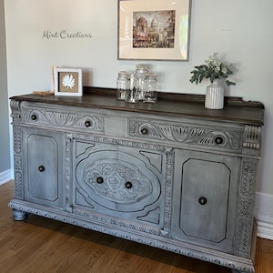 Peut inclure: Un grand buffet rectangulaire en bois avec une finition grise patinée et des détails sculptés complexes sur les façades des tiroirs et les portes centrales. Il possède un plateau supérieur brun foncé, des boutons ronds et des pieds boules.
