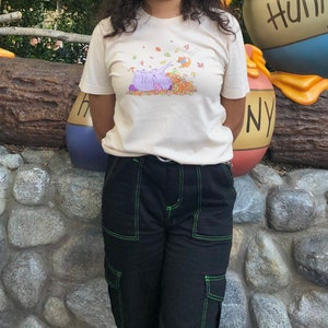 Könnte beinhalten: Ein cremefarbenes T-Shirt mit einem Aufdruck von zwei lila Elefanten mit einem gelben und orangefarbenen Blattdesign. Das Shirt hat kurze Ärmel und einen Rundhalsausschnitt.