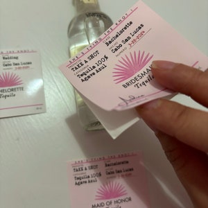 May include: A pink and white label with the text "She's Tying the Knot" and "Bachelorette" with a pink sun design. The label is attached to a clear glass bottle of tequila. The text "Take a Shot Tequila 100% Agave Azul" is printed on the label.
