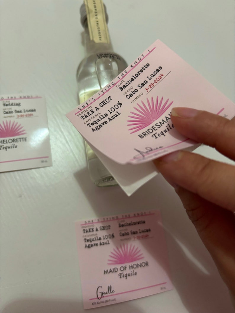 May include: A pink and white label with the text "She's Tying the Knot" and "Bachelorette" with a pink sun design. The label is attached to a clear glass bottle of tequila. The text "Take a Shot Tequila 100% Agave Azul" is printed on the label.