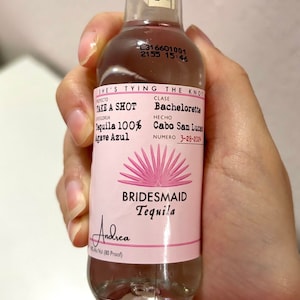 May include: A pink and white bottle of Bridesmaid Tequila with the text "Take a Shot Tequila 100% Agave Azul" and "She's Tying the Knot Bachelorette Cabo San Lucas 3-28-2024" on the label.